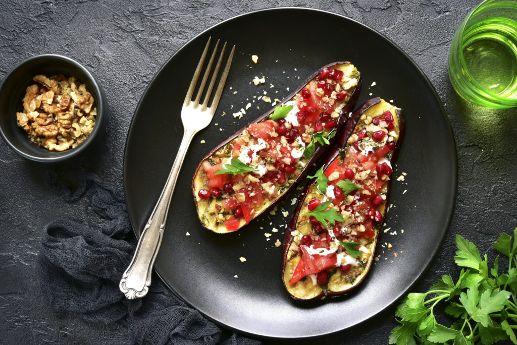 Grilled eggplant stuffed with tomatoes, nuts, pomegranate and yogurt dressing on a dark plate over black slate, stone or concrete background.Top view.